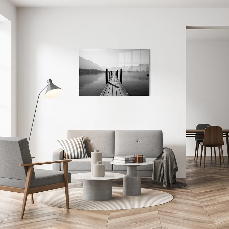 182074922_lifestyle A glass print hanging on a wall of a tranquil black and white wooden pier leading to a lake, with mountains silhouetted in the background.