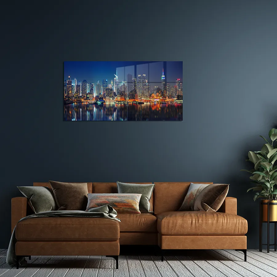 277637491_lifestyle A landscape glass print hanging a wall, picturing the skyline of Manhattan at night, with glowing lights reflected on the water.